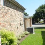 A side yard with green grass runs beside a brick house. There are small shrubs along the house, an air conditioning unit, and a wooden fence on the right. Trees and a paved street are visible in the background.