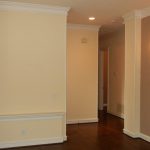 A room with cream-colored walls, dark wood floors, a wall-mounted light fixture, crown molding, and a large window with blinds letting in natural light. An open hallway is visible in the background.