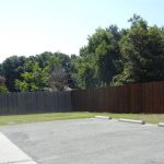 A small, empty parking lot with three concrete wheel stops, bordered by a tall wooden fence and trees under a clear blue sky.
