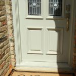 A tan doormat with the word “Welcome” in cursive sits in front of a cream-colored door with decorative glass panels. Part of the door is in sunlight and part is in shadow. The door is bordered by brick walls.