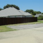 A residential house with a gray roof is behind a tall wooden fence. In the foreground, there is an empty parking lot with one black pickup truck parked. Trees and bushes surround the area.