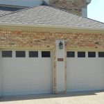 Two beige double garage doors on a brick house, with a black outdoor lantern and house number "6588" mounted between the doors. There is a concrete driveway and green grass along the edge.