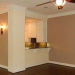 An empty living room with tan and beige walls, dark wood flooring, wall sconces, ceiling fan, and an open view into a kitchen with white cabinets and a black countertop.