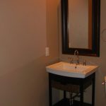 A small bathroom with beige walls, a rectangular mirror above a white sink with a black vanity, and a white toilet to the right. A towel ring is mounted on the wall next to the sink.