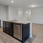 A modern, unfurnished kitchen with dark cabinets, granite countertops, a center island with a sink and dishwasher, and an open floor plan leading to a bright living area with wood-style flooring.