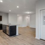 Modern kitchen with dark cabinets, granite countertops, and stainless steel appliances opens to a spacious, light-filled living area with wood-style flooring. A white door with a window is on the right side of the image.