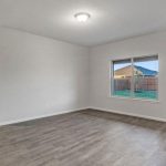 Unfurnished room with light gray walls, wood-look flooring, a ceiling light fixture, and a window showing a fenced backyard and neighboring houses. There is an open doorway on the left and overall natural lighting.