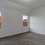 Empty room with light gray walls, two tall windows showing a fenced backyard with grass, and brown carpet flooring. Natural light enters from the windows, creating a bright atmosphere.