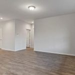 A spacious, empty room with light gray walls, wood-look flooring, a ceiling light fixture, and large windows on the right letting in natural light. Multiple doorways lead to other areas of the home.