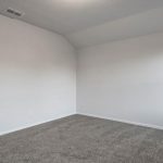 Empty room with light gray carpet, white walls, a door to the left, and a window to the right letting in natural light. The ceiling is sloped, and the room is clean and unfurnished.