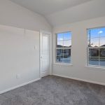 Empty room with light gray carpet, white walls, and two windows letting in natural light. A white door is between the windows, and houses can be seen outside. The room has a simple, clean appearance.