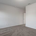 Empty room with light gray walls, a white ceiling, and brown carpeted flooring. There is a window on the left wall and a white door slightly open on the right side. The room is well-lit with natural light.