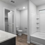 Modern bathroom with a double sink vanity, white countertops, a toilet, and a white bathtub/shower combo with built-in shelves. The space has wood-look flooring and white walls.