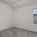 Empty room with white walls, gray carpeted floor, a window showing a view of an exterior brick wall, sloped ceiling, and electrical outlets visible along the walls.