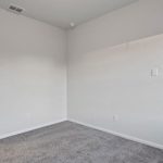 Empty bedroom with light gray carpet, white walls, and two white doors—one closed and one open, revealing a glimpse of an adjacent bathroom. Ceiling vent and smoke detector are also visible.