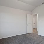 Empty bedroom with light gray walls, white ceiling, gray carpet, and a large window on the left. An open door leads to a hallway with wood-look flooring. Room is brightly lit by natural light.