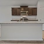 Modern kitchen with granite countertops, dark wood cabinets, a built-in microwave, and a stainless steel oven. The kitchen island has a sink and faucet, and the floor is light wood. Walls are light-colored, creating a bright space.