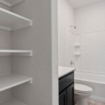 A bathroom with white shelves on the left, a white bathtub with a shower and built-in shelves, a toilet, and a dark-colored vanity with a white countertop on the right. The walls are white.
