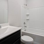 A clean, modern bathroom with a white bathtub and shower combo, white tiled walls, a toilet, and a white sink with a dark cabinet beneath and a large mirror above.