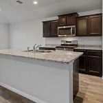 Modern kitchen with a large granite island, dark wooden cabinets, stainless steel appliances, wood flooring, and a large window letting in natural light.