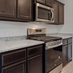 Modern kitchen with dark wood cabinets, granite countertops, a stainless steel gas stove and oven, built-in microwave, light gray walls, and a white door to the right.