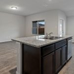 A modern, empty kitchen with a large granite island and sink, dark cabinets, wood-look flooring, and several windows letting in natural light. The open layout connects to a spacious living area.